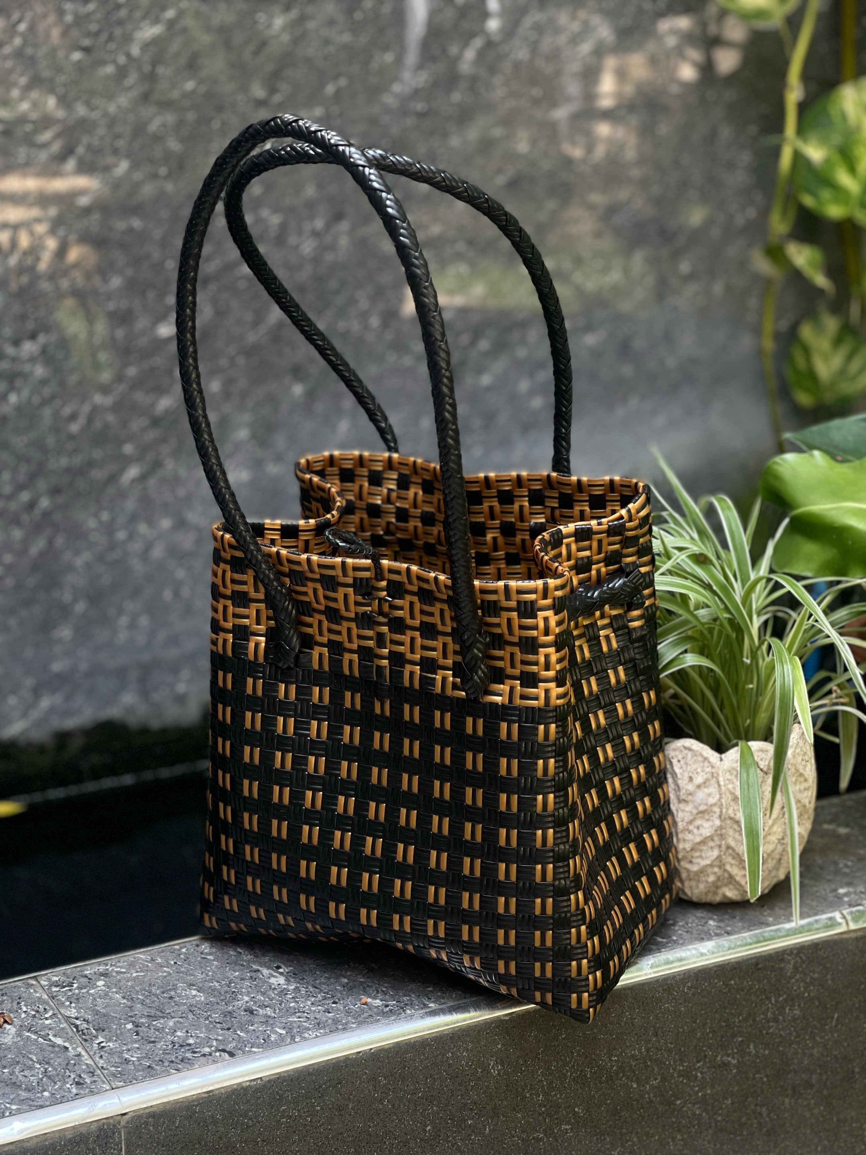 A medium-sized handbag with black straps and gold body, placed on a surface with a plant in the background.